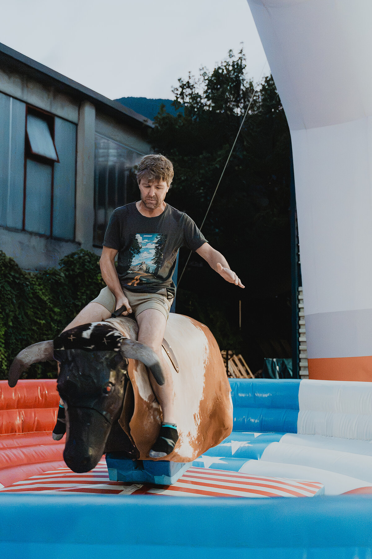 Ein Mann reitet auf einem mechanischen Bullen in einem aufblasbaren Rodeo-Bereich, der aus bunten Elementen besteht. Der Hintergrund zeigt ein Gebäude und Bäume.