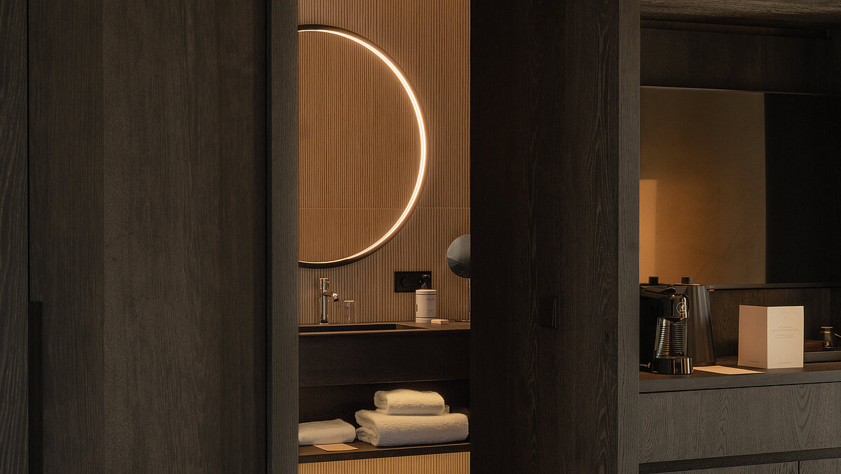 Ein modernes Hotelzimmer mit dunklem Holzinterieur. Im Hintergrund ist ein Badezimmer mit beleuchtetem runden Spiegel und weißen Handtüchern zu sehen. Im Vordergrund steht ein weißes Bett.
