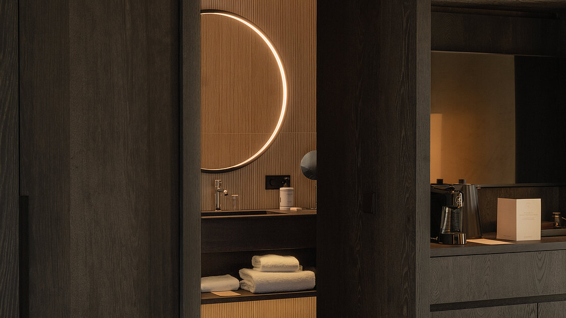 Ein modernes Hotelzimmer mit dunklem Holzinterieur. Im Hintergrund ist ein Badezimmer mit beleuchtetem runden Spiegel und weißen Handtüchern zu sehen. Im Vordergrund steht ein weißes Bett.
