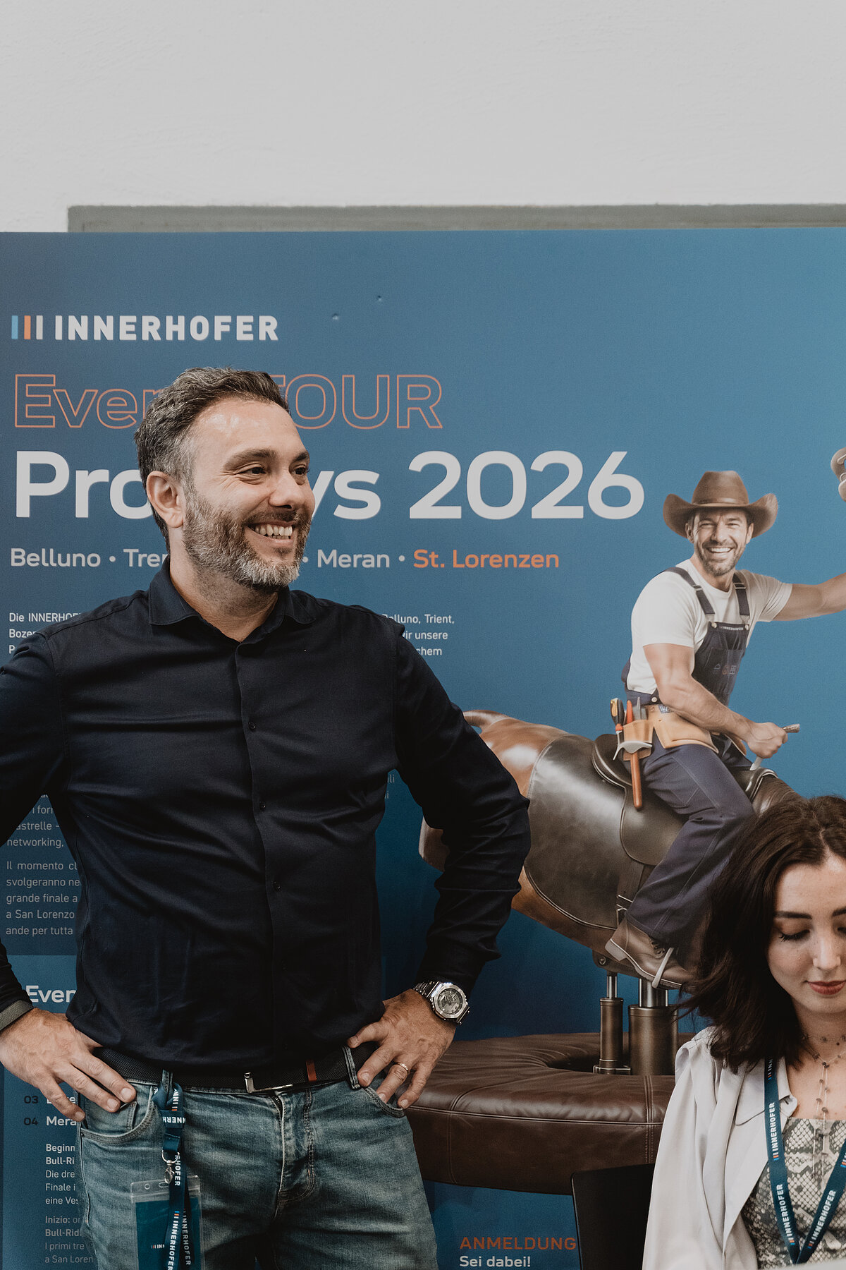 Ein Mann steht lächelnd vor einem blauen Werbeplakat mit der Aufschrift "Eventour ProDays 2026". Rechts sieht man eine Frau, die nach unten schaut. Im Hintergrund ist ein Bild von einem lachenden Mann in Arbeitskleidung, der auf einem Pferd reitet.