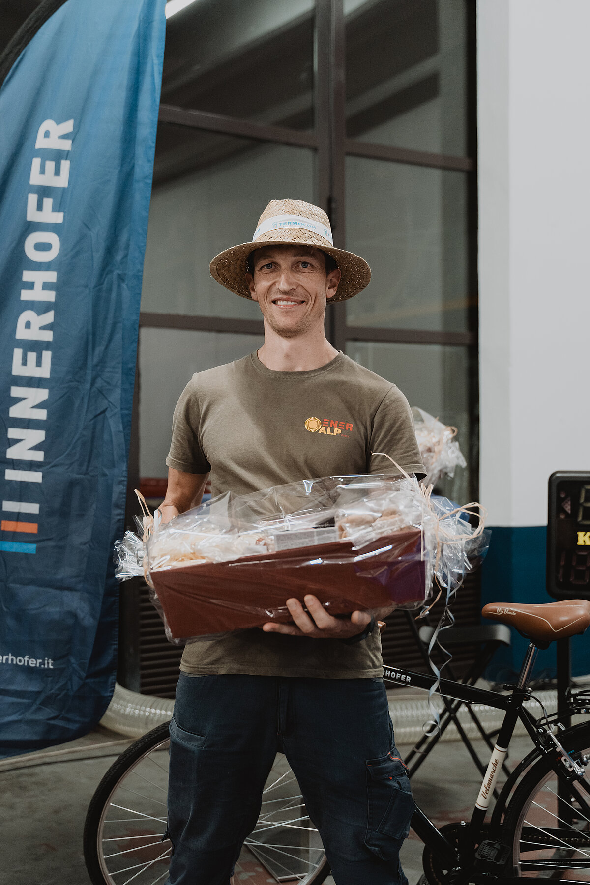 Ein Mann mit Strohhut hält einen Geschenkkorb und steht vor einem Fahrrad. Links neben ihm ist eine blaue Fahne mit der Aufschrift "INNERHOFER" zu sehen.