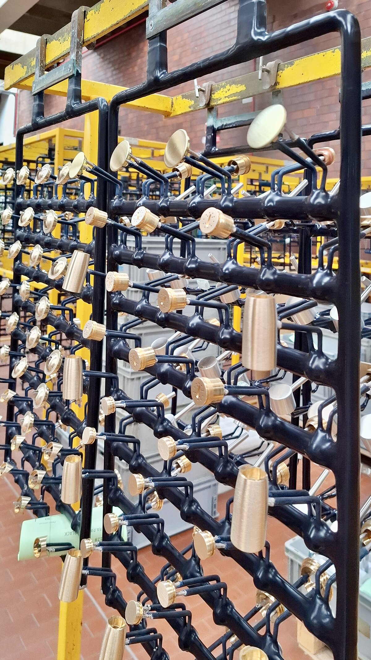 Ein Metallgestell mit vielen goldfarbenen Wasserhahn-Teilen, die an schwarzen Halterungen befestigt sind. Das Gestell steht in einer Fabrikhalle mit ziegelrotem Boden und gelben Trägern im Hintergrund.