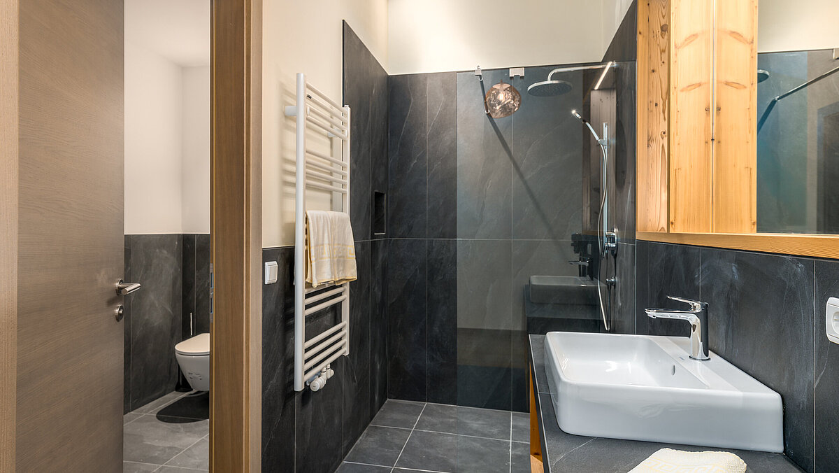 Bagno moderno con piastrelle scure, lavabo bianco con rubinetto cromato, e specchio ampio. A destra, asciugamani piegati su un ripiano. A sinistra, porta aperta che conduce a un gabinetto. Atmosfera luminosa e ordinata.