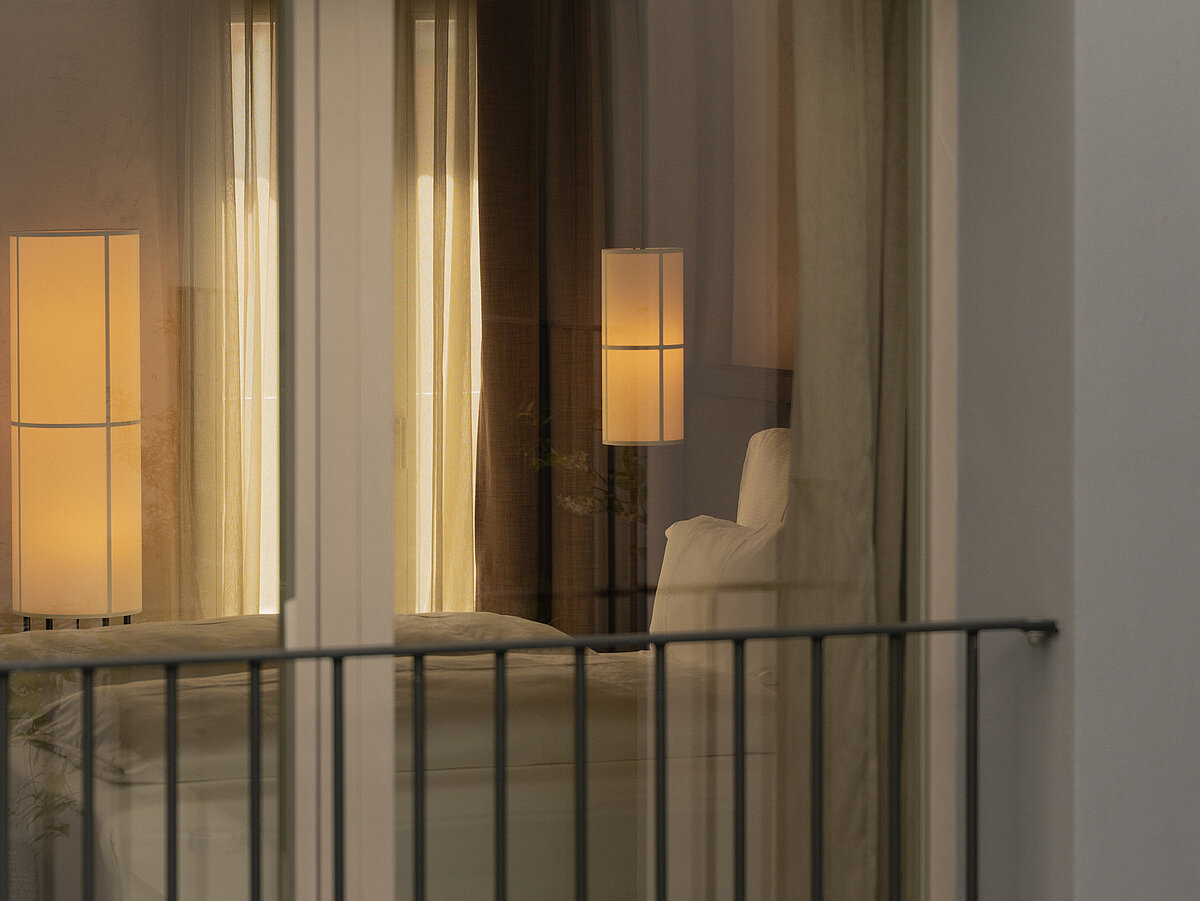 Fensteransicht eines gemütlich beleuchteten Schlafzimmers mit einem Bett und einer Stehlampe. Ein modernes Balkongeländer ist im Vordergrund zu sehen.