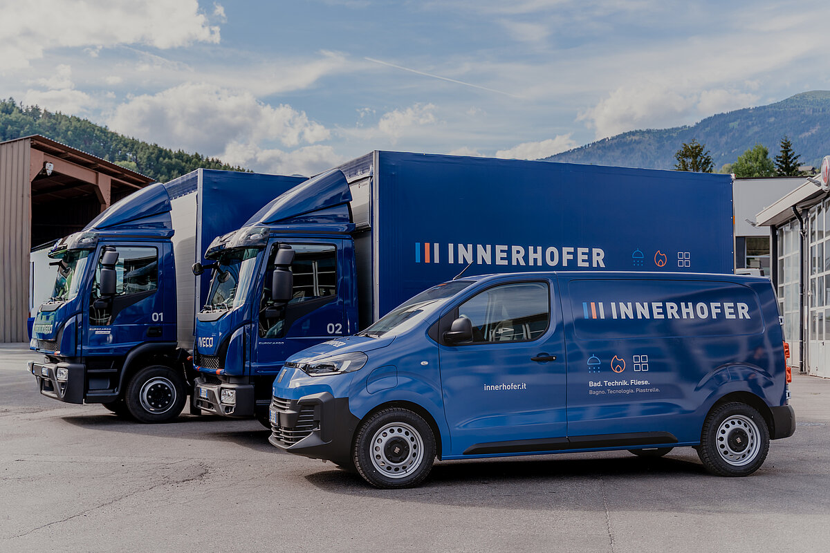 Drei blaue Lieferfahrzeuge der Marke Innerhofer stehen nebeneinander auf einem Parkplatz, umgeben von Bergen. Die Fahrzeuge sind mit dem Firmenlogo und Schriftzügen versehen. Der Himmel ist leicht bewölkt.