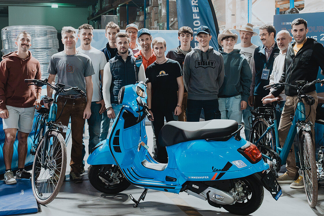 Eine Gruppe von Menschen steht in einem Lager vor einem blauen Motorroller und mehreren Fahrrädern. Im Hintergrund sind Regale mit Kartons zu sehen. Die Gruppe posiert für ein Foto neben einer Werbebannerflagge.