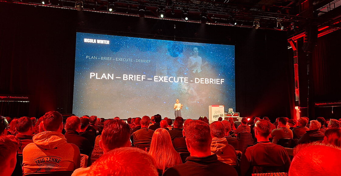 Eine Person gibt eine Präsentation vor einer großen Zuhörerschaft. Auf der Projektionsfläche hinter ihnen steht „PLAN - BRIEF - EXECUTE - DEBRIEF".