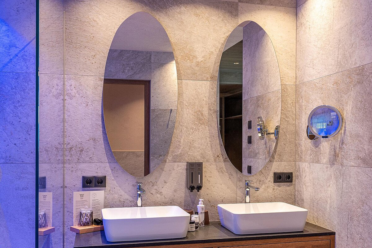 Modernes Badezimmer mit zwei ovalen Spiegeln über einem Holzbalken mit zwei rechteckigen Waschbecken. Der Raum ist in einer warmen Beleuchtung und blauen Akzentlichtern gestaltet, mit grauen Fliesen an Wänden und Boden.