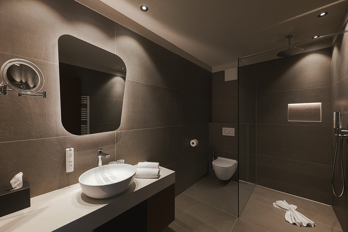 Modernes Badezimmer mit großer begehbarer Dusche, Wandtoilette und Waschtisch mit rundem Waschbecken. Dezente Beleuchtung und dunkle Fliesen prägen das stilvolle Design. Handtücher und dekorative Elemente ergänzen die Einrichtung.