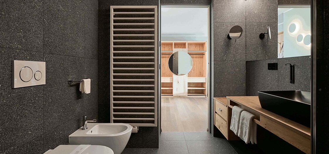 Ein modernes Badezimmer mit dunklen Fliesen, einem Waschbecken mit schwarzer Armatur, einem Bidet, einer Toilette und einem Handtuchwärmer. Im Hintergrund befindet sich ein Ankleideraum mit Holzregalen und einem runden Spiegel.