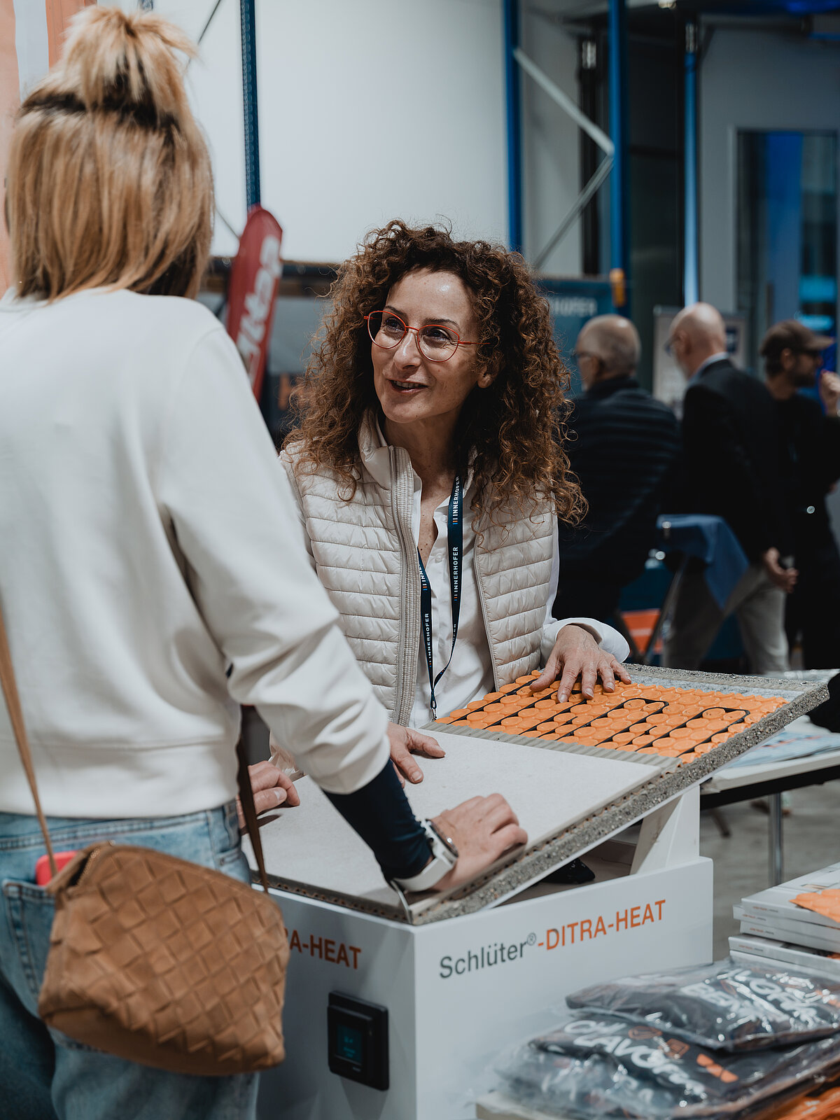 Zwei Frauen sprechen an einem Messestand über ein Produkt namens "Schlüter-DITRA-HEAT". Die Frau auf der rechten Seite trägt eine Brille und lächelt, während sie die Installation von Heizungsmatten erklärt. Im Hintergrund sind weitere Messebesucher zu sehen.