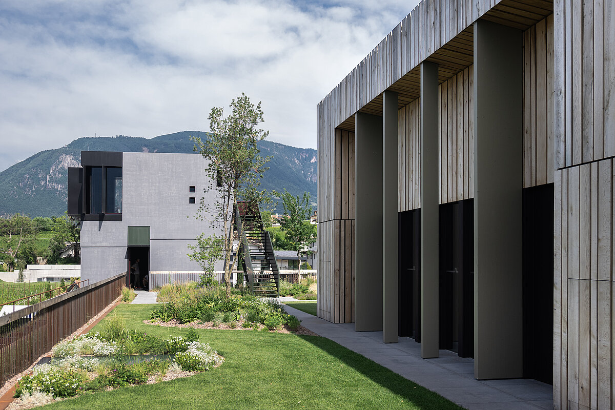 Moderne Architektur mit klaren Linien und hölzernen Elementen, umgeben von grünen Landschaften und Gärten. Im Hintergrund sind Berge unter einem teilweise bewölkten Himmel zu sehen.