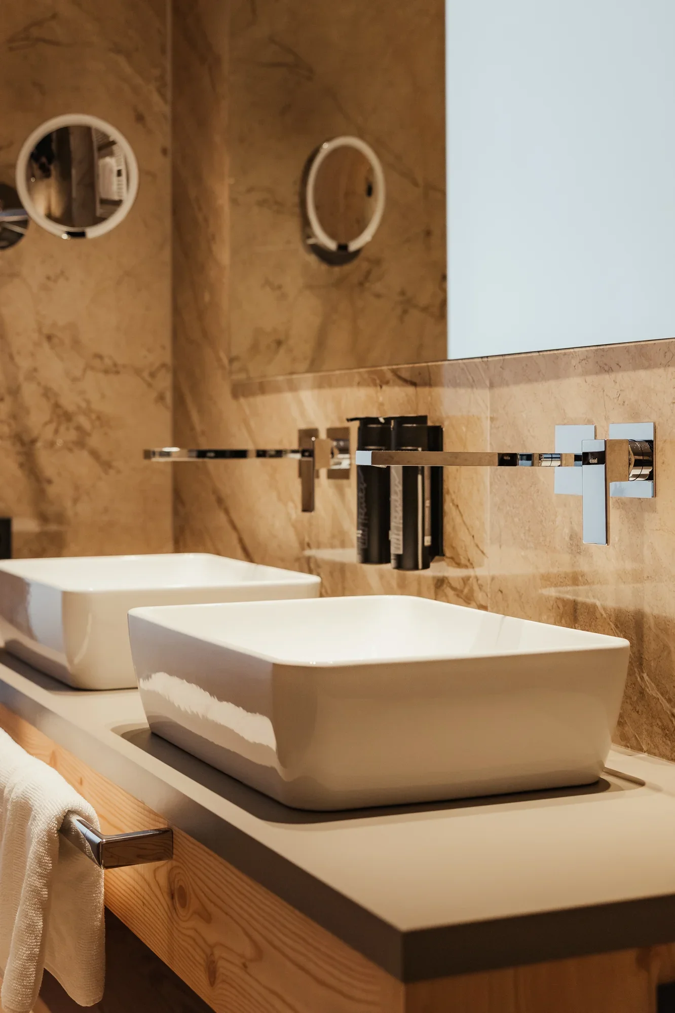 Modernes Badezimmer mit zwei rechteckigen Aufsatzbecken auf einem Waschtisch aus Holz. An der Wand befinden sich zwei runde Spiegel und integrierte Wasserhähne. Die Wände sind aus Marmor. Ein Handtuch hängt am Waschtisch.
