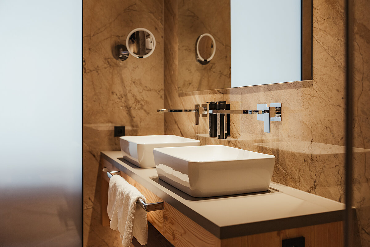 Modernes Badezimmer mit zwei rechteckigen Aufsatzbecken auf einem Waschtisch aus Holz. An der Wand befinden sich zwei runde Spiegel und integrierte Wasserhähne. Die Wände sind aus Marmor. Ein Handtuch hängt am Waschtisch.