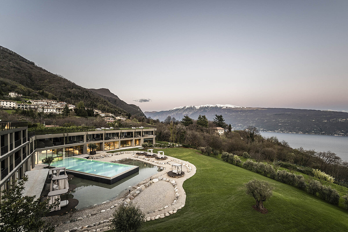 Ein modernes Gebäude mit großen Fenstern steht neben einem rechteckigen Swimmingpool, eingebettet in eine gepflegte Grünfläche. Im Hintergrund erstreckt sich ein See, umgeben von bewaldeten Hügeln und Bergen.