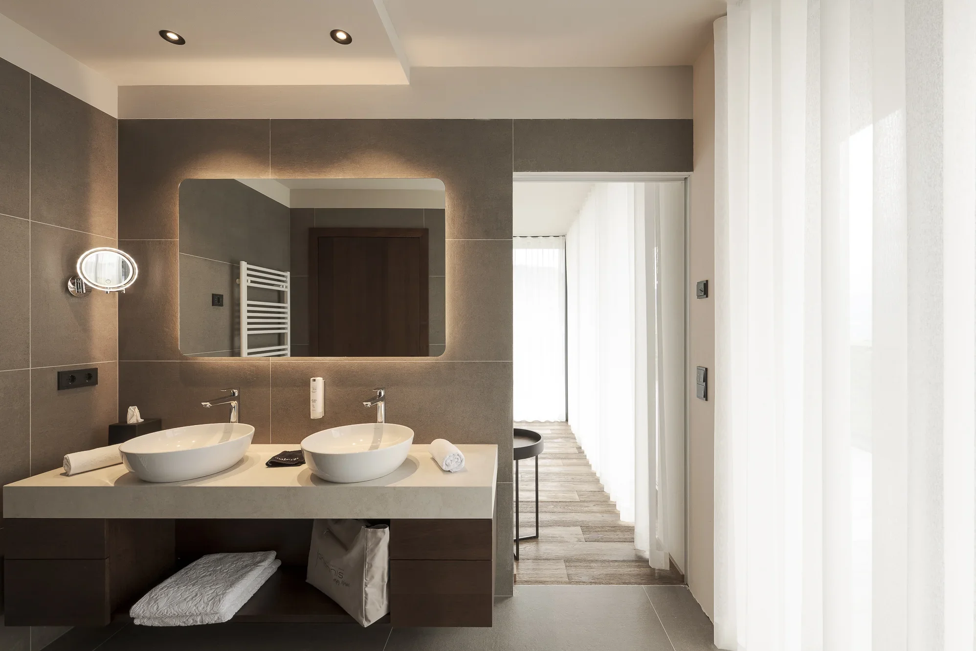 Ein modernes Badezimmer mit Doppelwaschbecken, großem Spiegel und schlichter Dekoration. Die Raumgestaltung ist minimalistisch, mit grauen Wänden und hellen Bodenfliesen. Ein Handtuch liegt ordentlich gefaltet neben den Waschbecken.