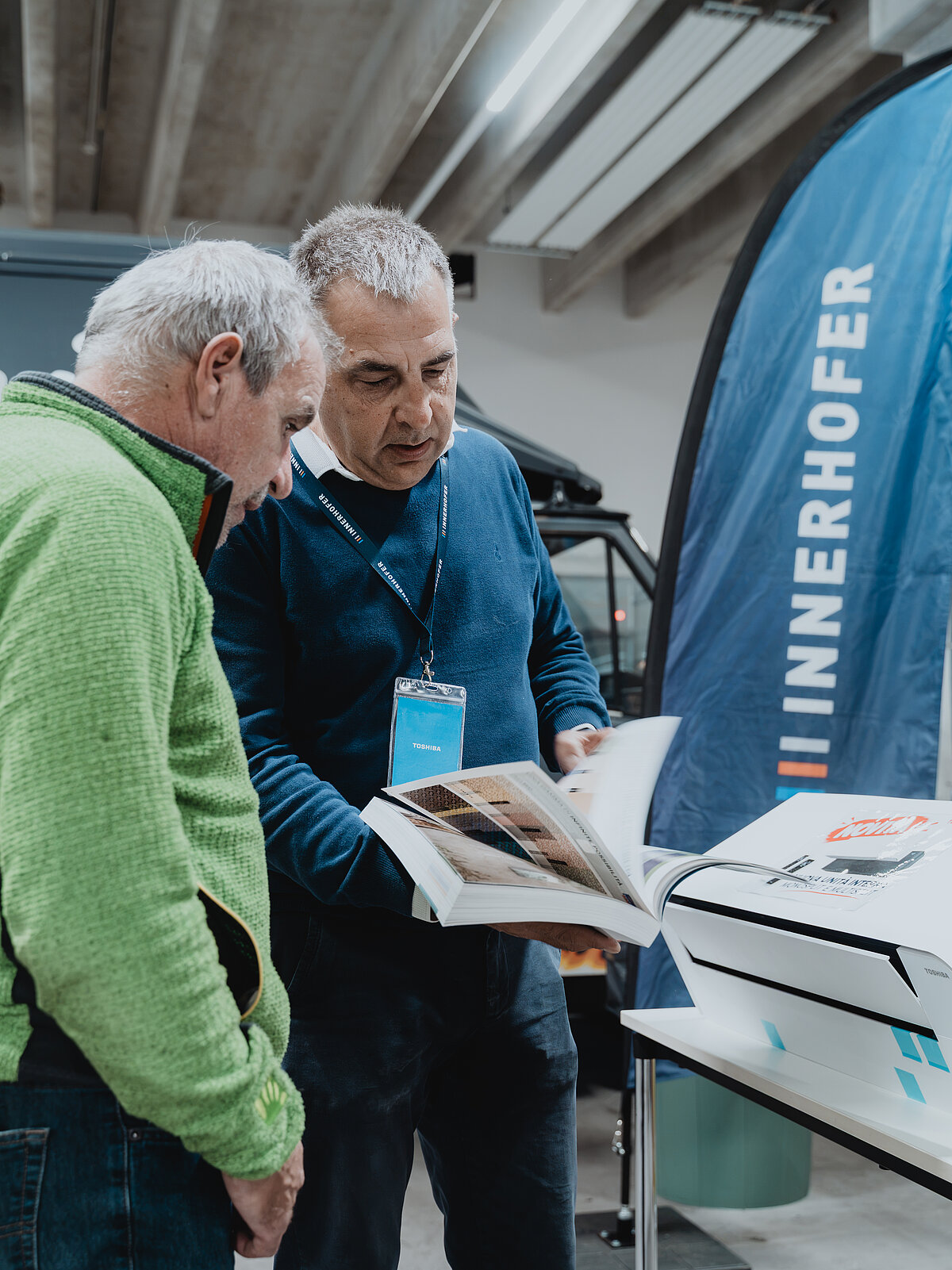 Zwei Männer schauen sich gemeinsam ein offenes Buch oder Katalog an. Einer trägt einen grünen Pullover, der andere einen blauen. Im Hintergrund ist ein Werbebanner mit dem Schriftzug "INNERHOFER" sichtbar. Die Umgebung wirkt wie eine Messe oder ein Ausstellungsraum.