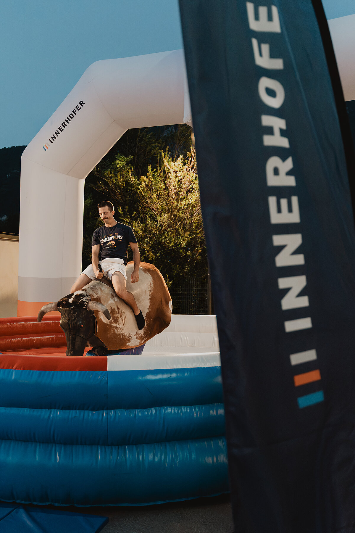 Ein junger Mann reitet auf einem mechanischen Bullen innerhalb eines aufgeblasenen Rings. Im Vordergrund ist ein Banner mit der Aufschrift „INNERHOFER“ zu sehen. Im Hintergrund sind Bäume und ein Teil eines Gebäudes unter dem Abendhimmel.