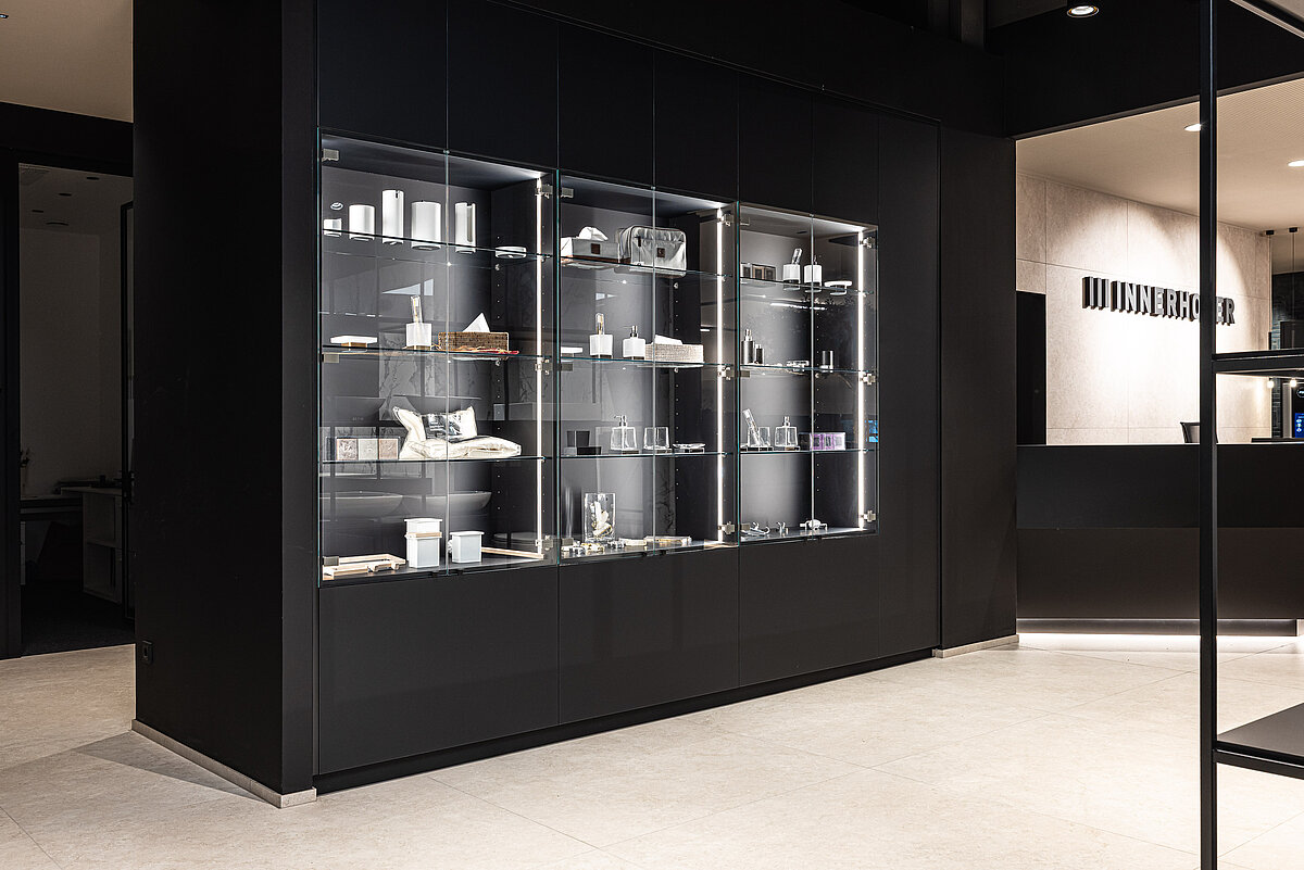 Ein moderner Innenraum mit einem großen, beleuchteten Vitrinenschrank voller weißer Tassen und kleinerer Gegenstände. Neben der Vitrine befindet sich ein Empfangstresen mit einem Schild darüber. Der Raum hat einen schlichten, minimalistischen Stil.