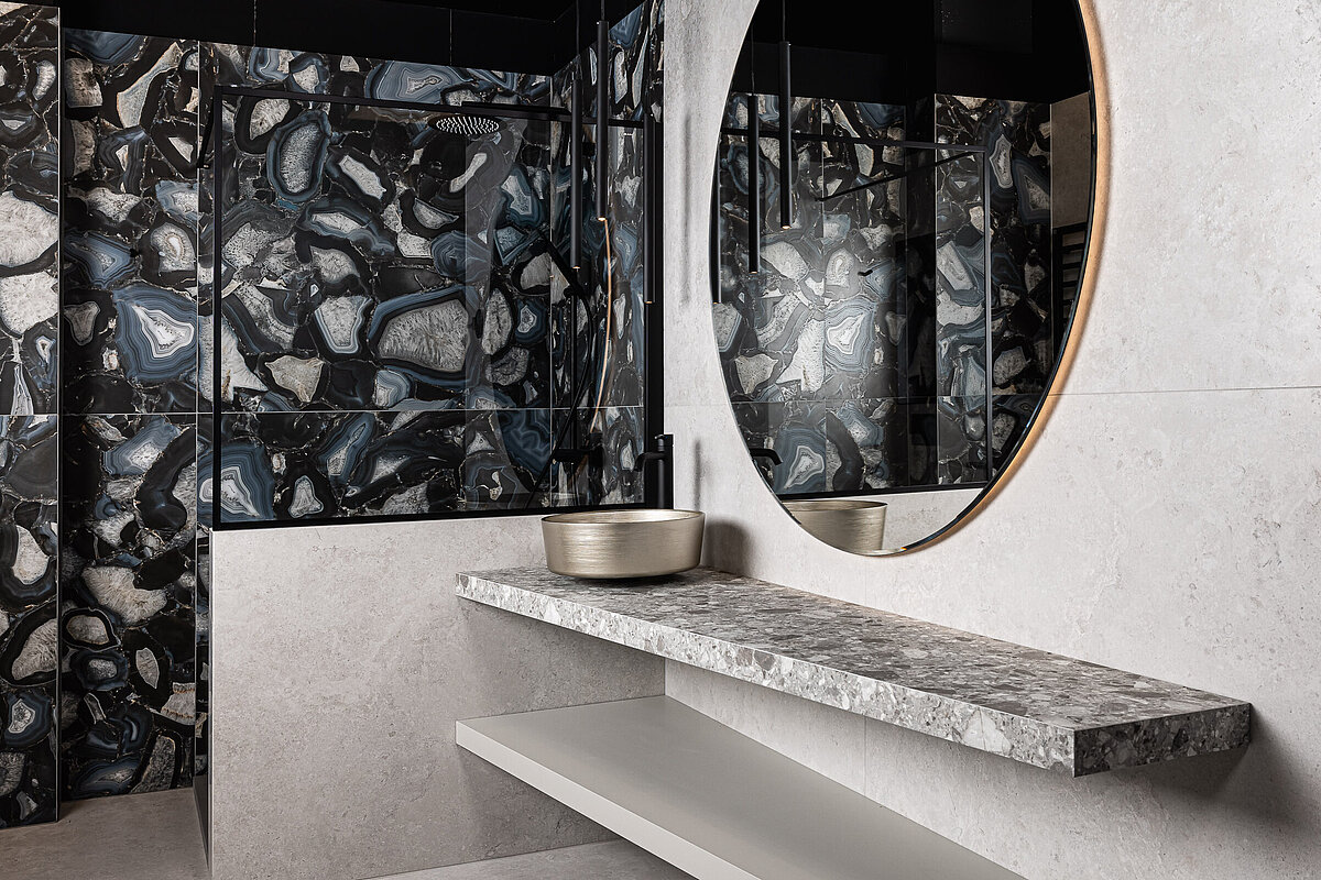 Modernes Badezimmer mit großem, rundem Spiegel und schwarz-grauen Fliesen mit abstraktem Muster. Dunkle Decke, heller Waschtisch mit grau gemusterter Oberfläche und darauf eine moderne, runde Waschschüssel.