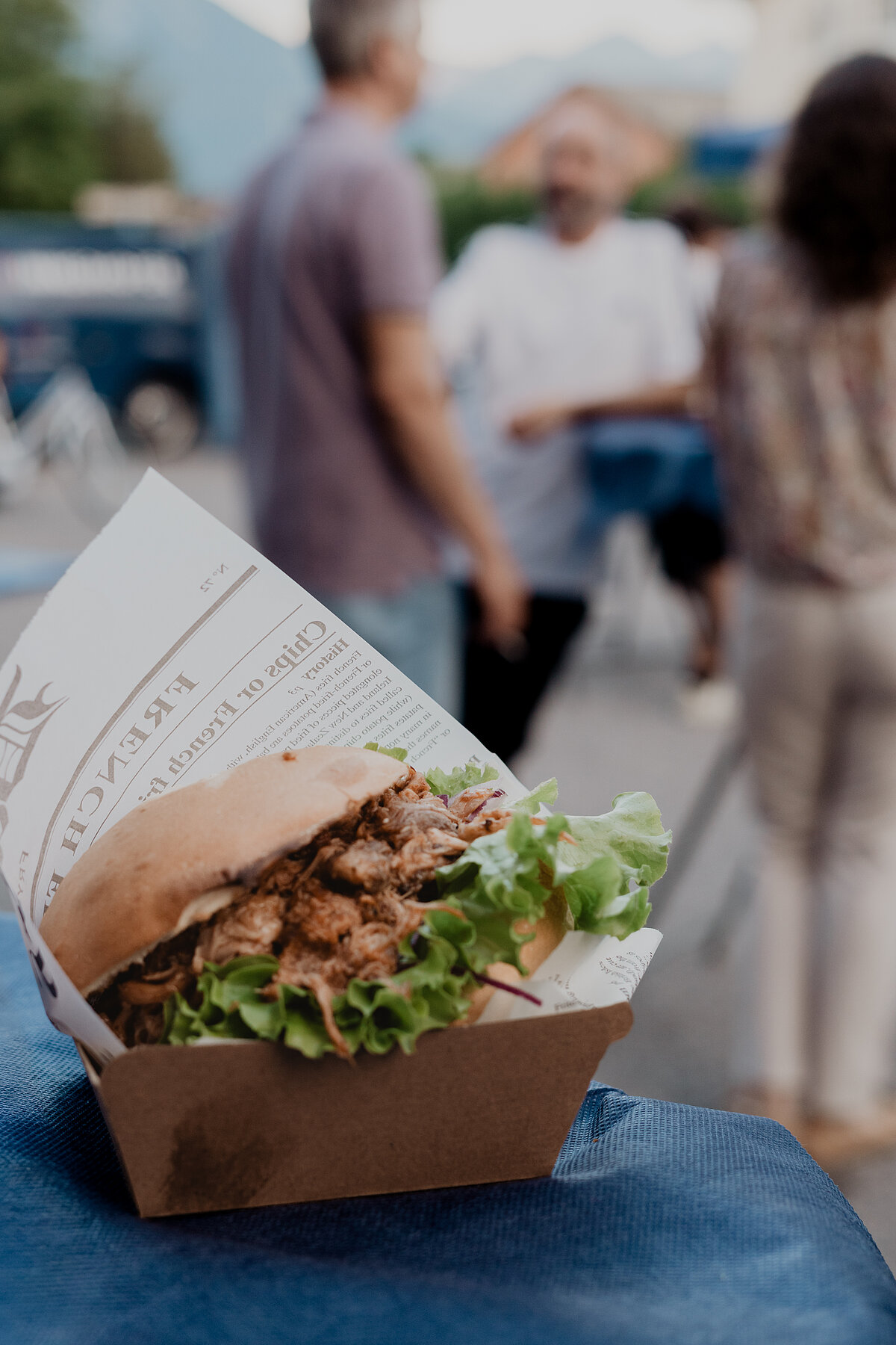 Ein Burger mit Salat und Fleisch in einer Pappschachtel wird auf einem Tisch präsentiert. Im Hintergrund sind verschwommene Personen bei einem Outdoor-Event zu sehen.