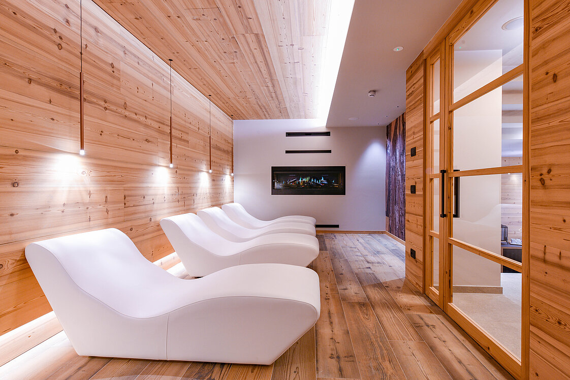 Eine moderne Entspannungslounge mit einer Reihe von weißen, ergonomischen Liegestühlen auf einem holzgetäfelten Boden. Die Wände und Decke sind ebenfalls aus Holz, mit sanfter Beleuchtung. Ein flacher Bildschirm ist an der Wand montiert, die Atmosphäre wirkt ruhig und erholsam.