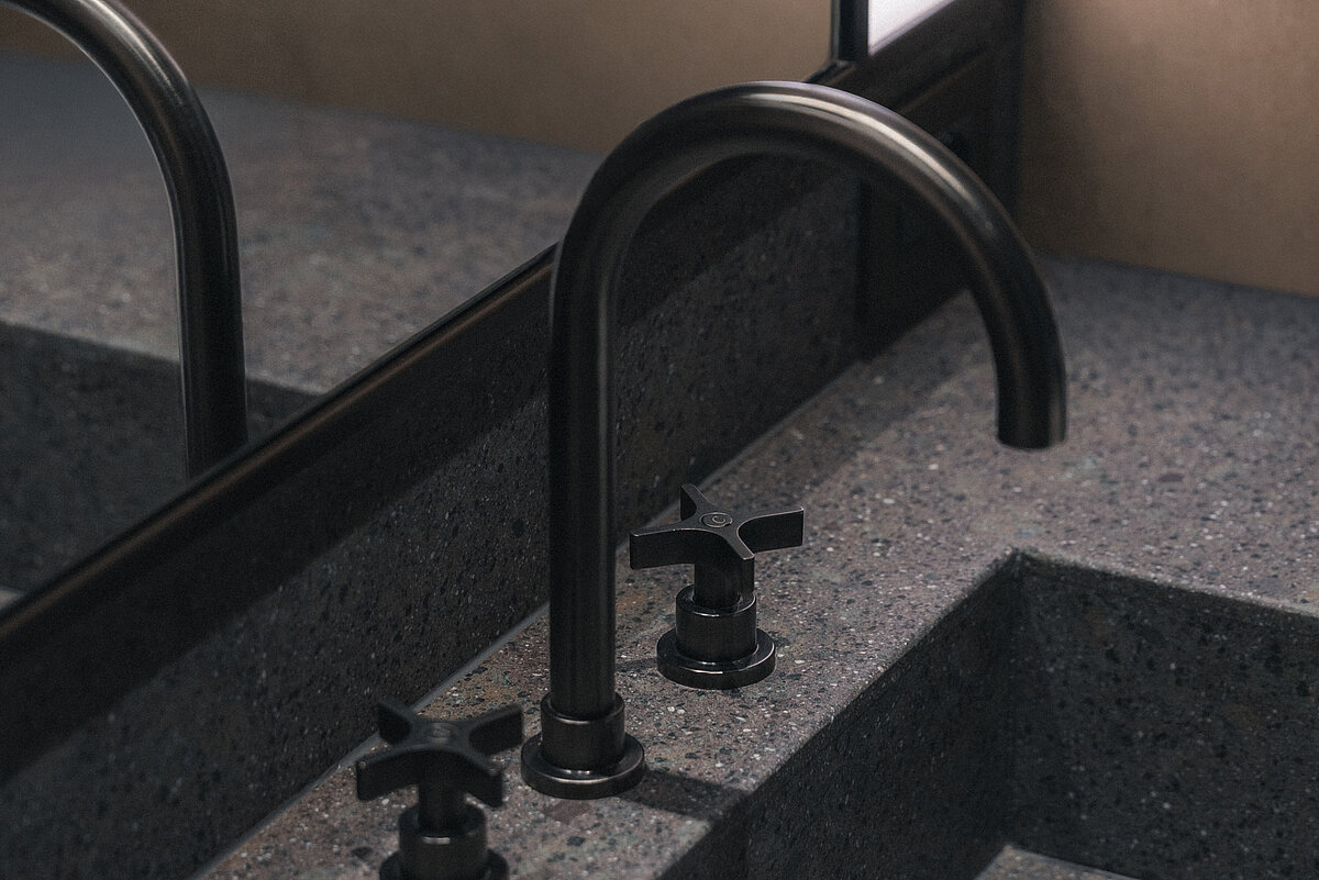 Ein modernes Waschbecken aus dunkelgrauem Stein mit einem schwarzen Wasserhahn und zwei Drehknöpfen. Im Hintergrund befindet sich ein Spiegel.