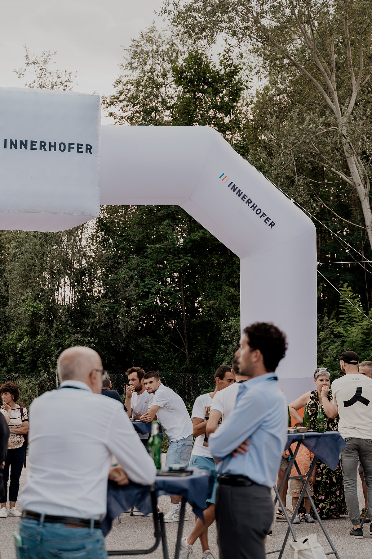 Menschen versammeln sich im Freien unter einem großen, aufblasbaren Eingangsbogen mit der Aufschrift „INNERHOFER“. Einige unterhalten sich an Stehtischen und andere stehen in kleinen Gruppen. Im Hintergrund sind Bäume zu sehen.