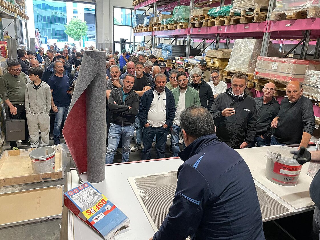 Eine große Gruppe von Menschen steht in einem Lagerhaus und beobachtet eine Person, die eine handwerkliche Demonstration an einem Tisch durchführt. Im Hintergrund sind Regale mit Waren zu sehen.
