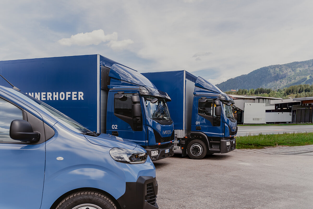Drei blaue Lastwagen mit der Aufschrift "INNERHOFER" parken nebeneinander auf einem Parkplatz. Im Hintergrund sind grüne Hügel und ein Gebäude zu sehen.