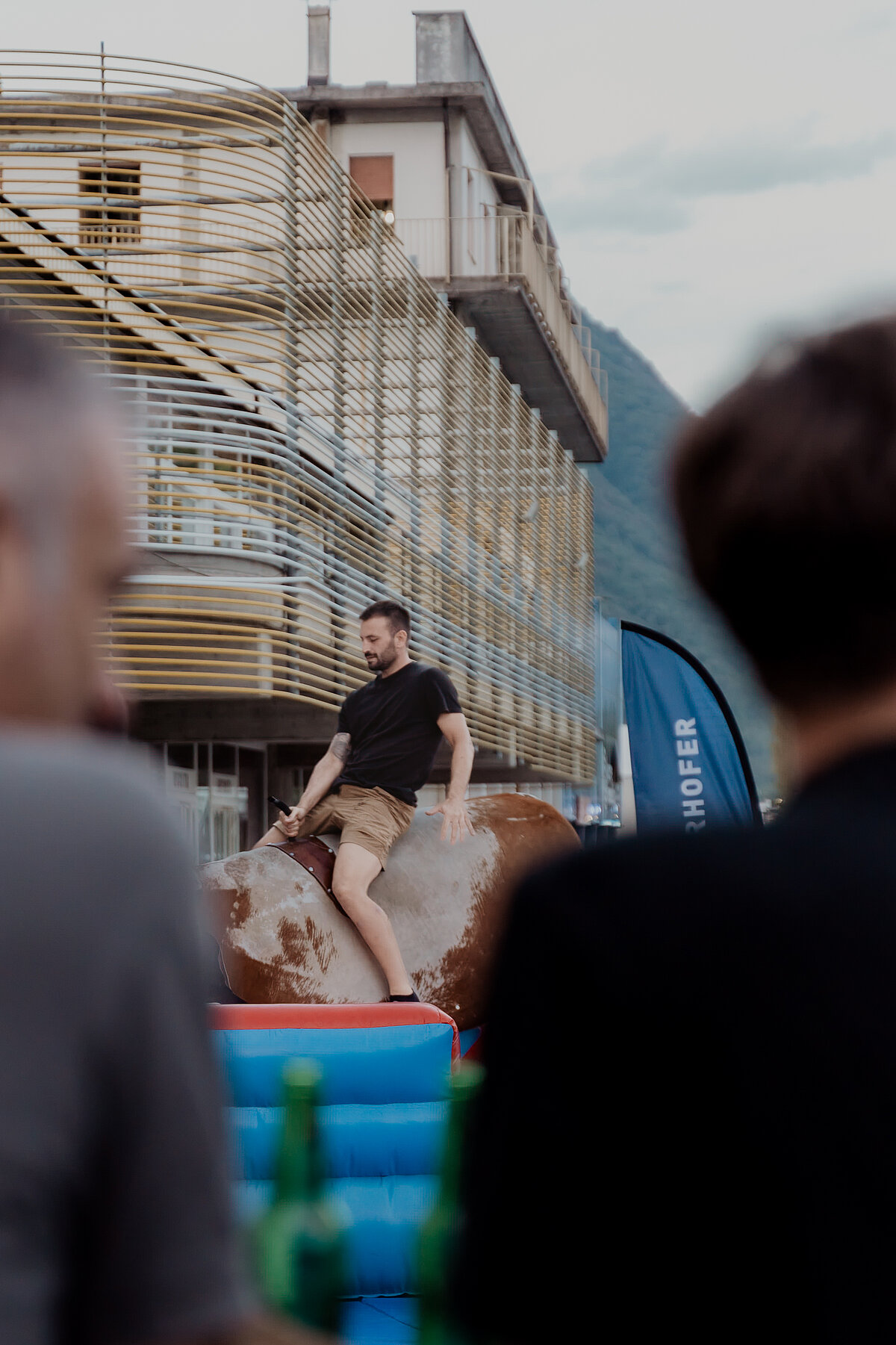 Ein Mann in Shorts und T-Shirt reitet auf einem mechanischen Bullen, während im Hintergrund moderne Gebäude und ein bewölkter Himmel zu sehen sind. Zuschauer mit Getränkeflaschen beobachten die Szene.