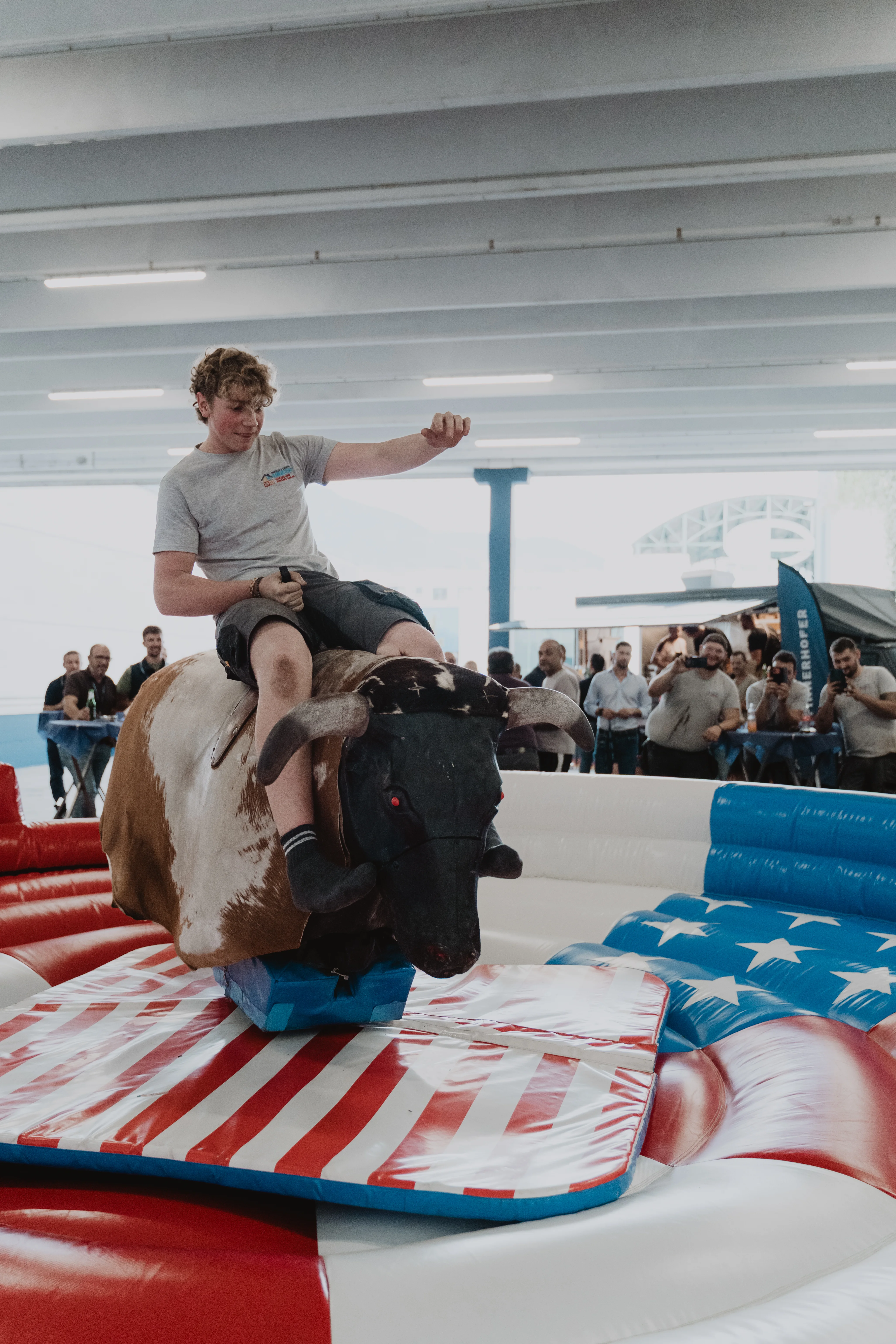 Ein Mann reitet auf einem mechanischen Bullen in einer Halle. Der Bulle steht auf einer Plattform mit einem Muster, das der amerikanischen Flagge ähnelt. Im Hintergrund sieht man eine Gruppe von Zuschauern, die das Geschehen beobachten.