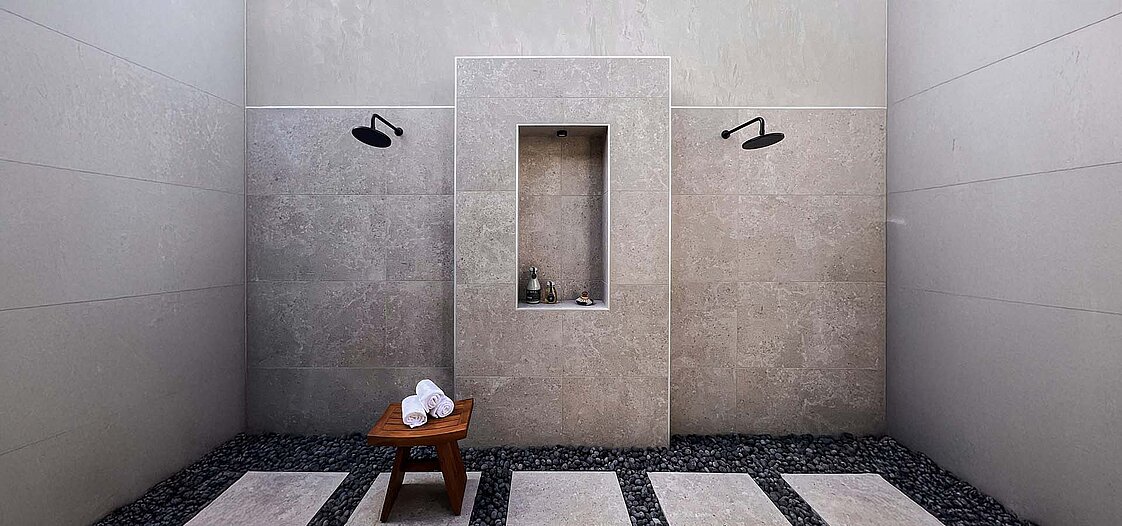 Eine moderne, minimalistische Dusche mit zwei Regenduschen an einer Steinwand. In der Mitte der Wand befindet sich eine kleine Nische mit Pflegeprodukten. Ein Holzhocker mit weißen Handtüchern steht auf einem gestreiften Boden aus Kieselsteinen.
