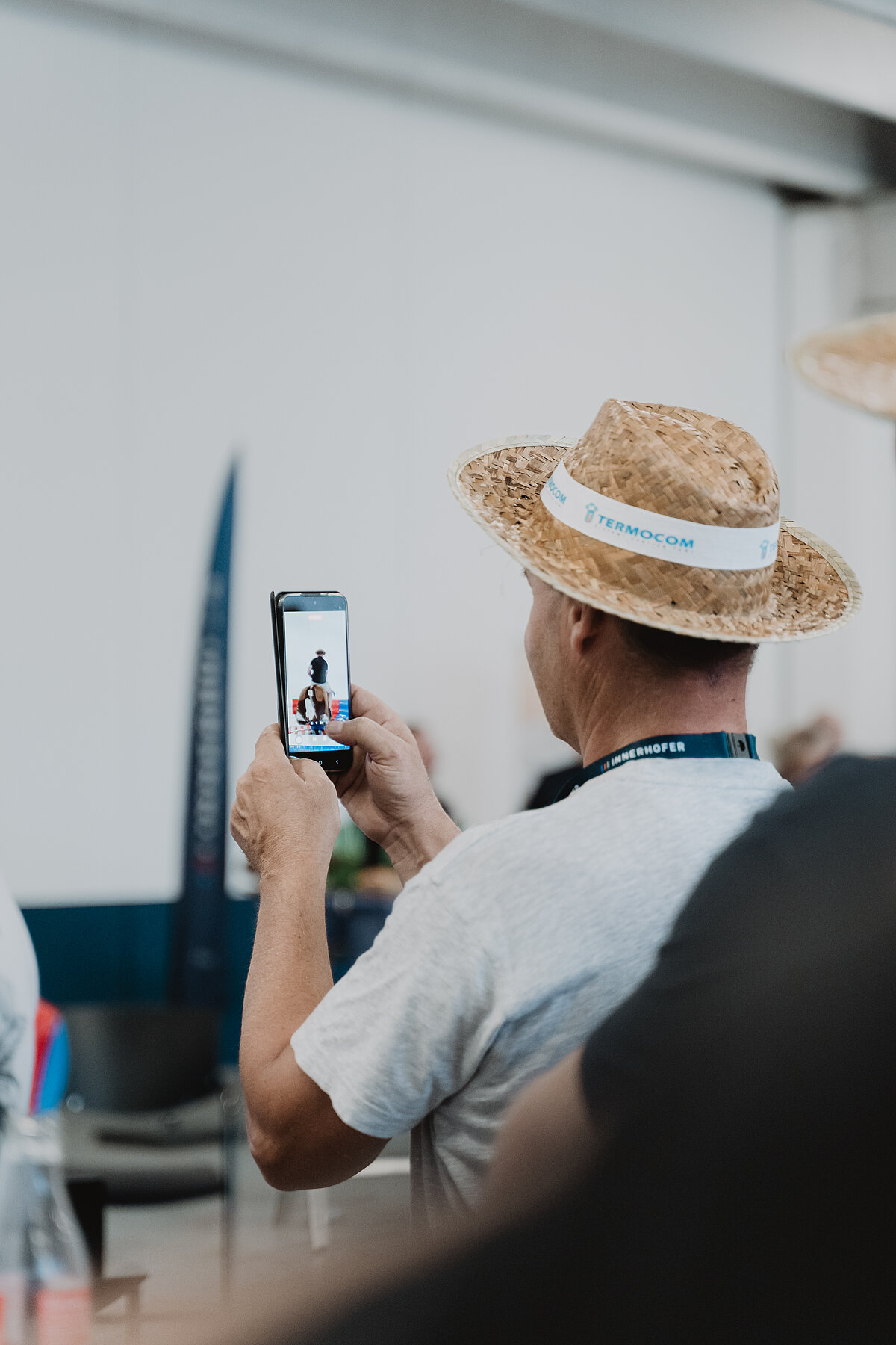 Ein Mann mit Strohhut fotografiert mit seinem Smartphone eine Person in einem Innenraum. Der Hut trägt ein Band mit der Aufschrift "Promocom". Der Hintergrund ist verschwommen, was den Fokus auf das Fotografieren legt.