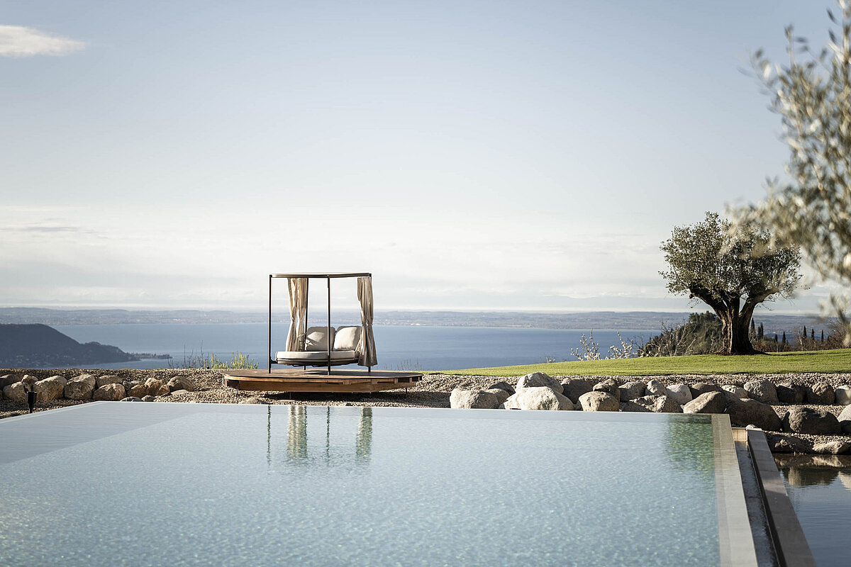 Ein Infinity-Pool mit Blick auf ein ruhiges Gewässer und daran angrenzend eine Liege mit Vorhängen. Im Hintergrund ist ein einzelner Baum und eine weite Landschaft zu sehen, die eine friedliche, entspannende Atmosphäre vermittelt.
