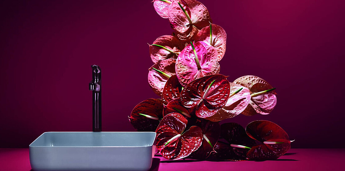 Ein lebendiges Arrangement aus rosa und roten Blumen steht auf einer Oberfläche vor einem dunkelrosa Hintergrund.