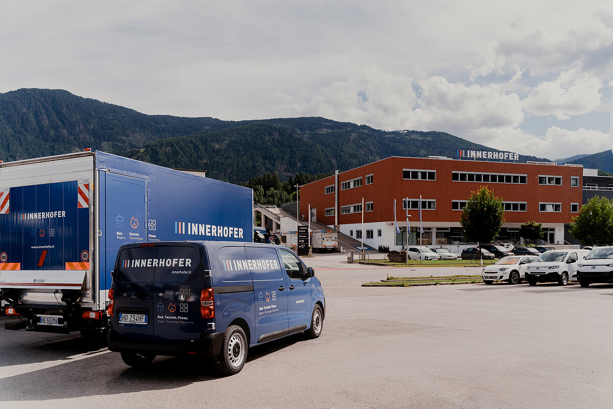 Zwei blaue Lieferfahrzeuge mit der Aufschrift "Innerhofer" stehen auf einem Parkplatz vor einem großflächigen, roten Geschäftsgebäude. Im Hintergrund sind bewaldete Hügel und ein bewölkter Himmel zu sehen.