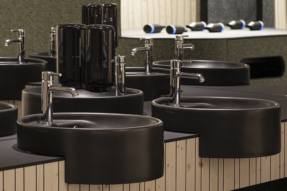 Schwarze runde Waschbecken mit silbernen Wasserhähnen auf einem gemusterten Holztresen in einem schicken Badezimmer. Im Hintergrund ist ein großer Spiegel und Beleuchtung zu sehen. Elegantes, modernes Design mit akzentuierten schwarzen Details.