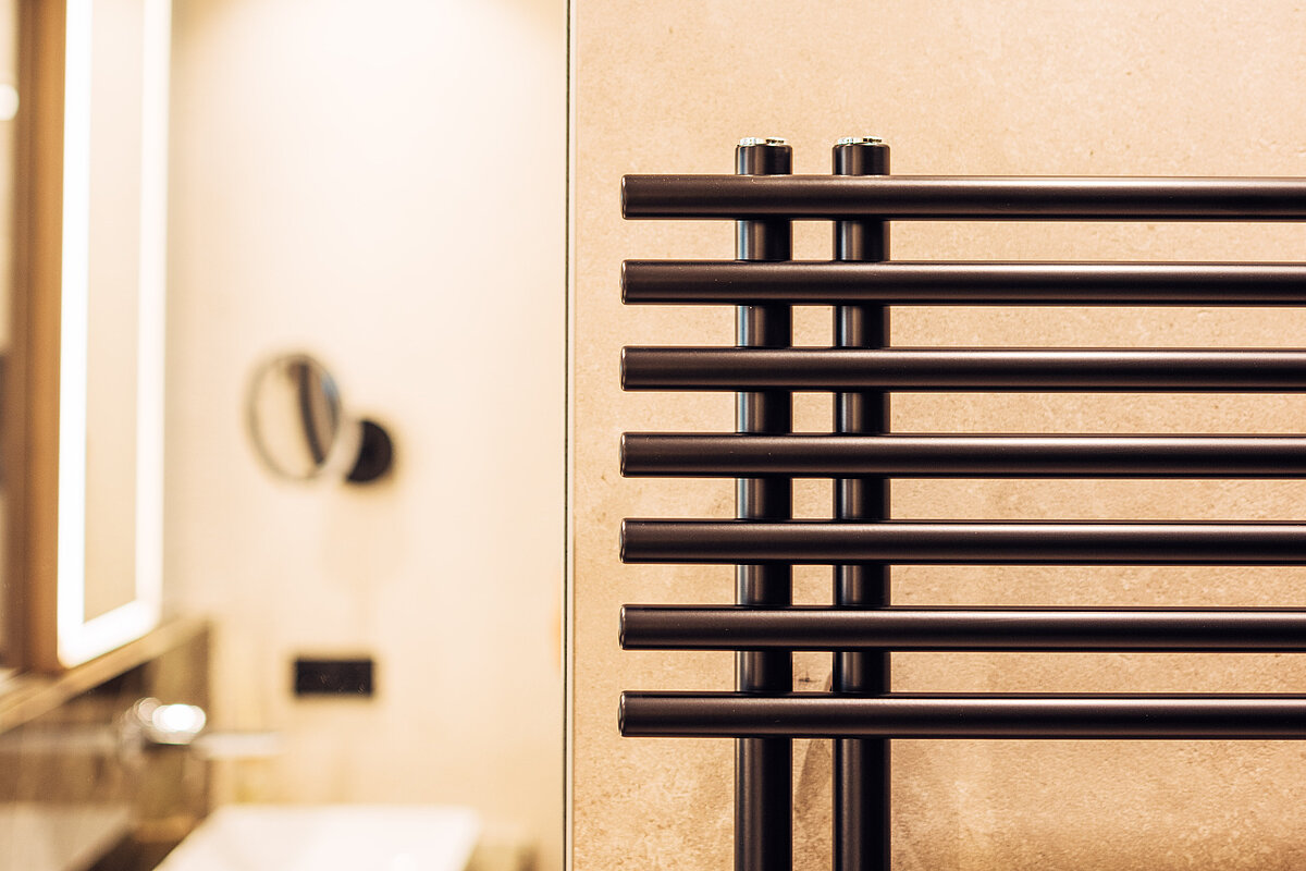 Ein elegantes Badezimmer mit einem modernen, schwarzen Handtuchheizkörper an der Wand. Im Hintergrund spiegelt sich ein Waschbecken und ein beleuchteter Spiegel auf beigen Fliesen.