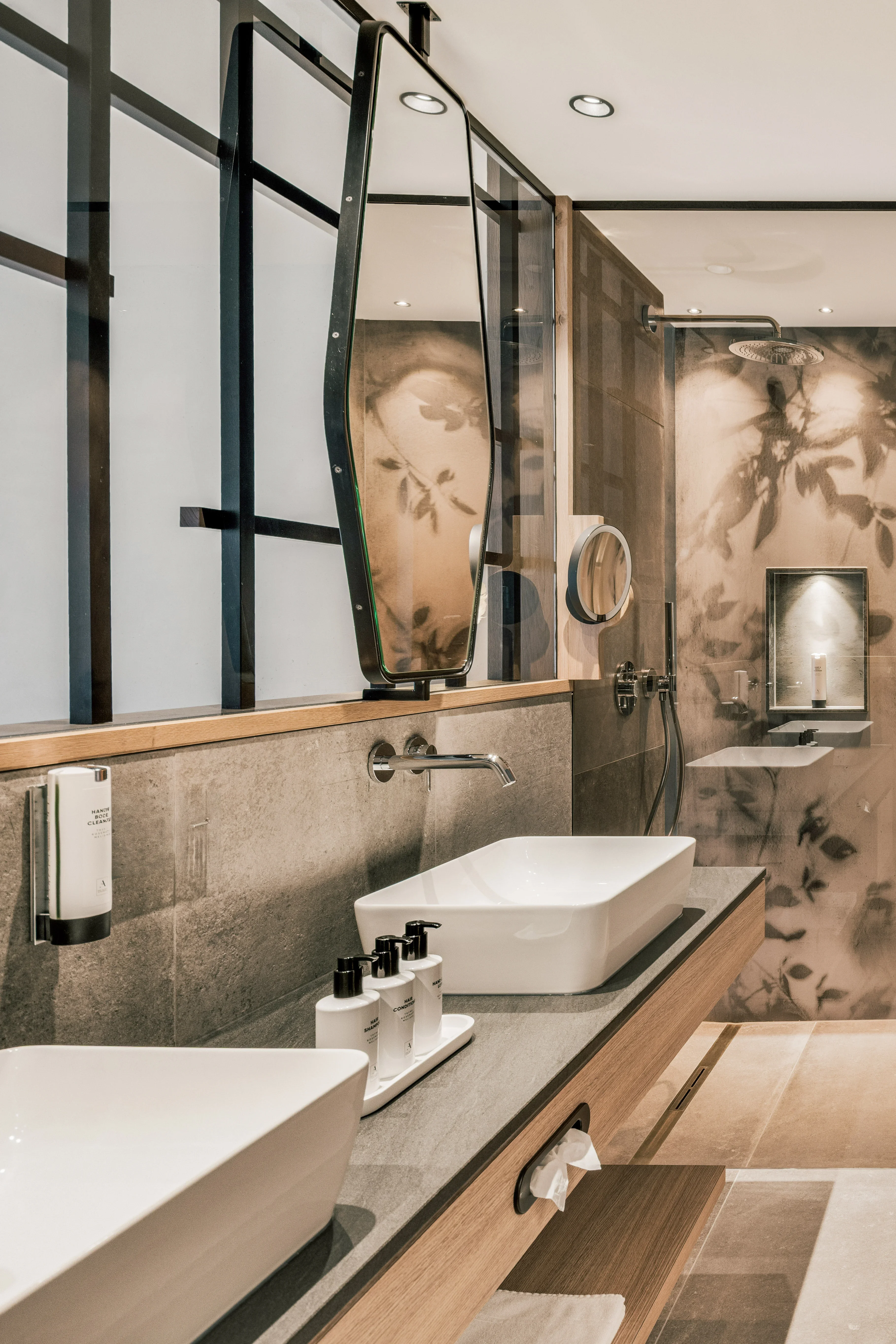 Modernes Badezimmer mit zwei Waschbecken auf einer Holzplatte, großem Spiegel, Glasdusche mit floralem Muster und Handtuchheizkörper. Beleuchtung schafft eine warme Atmosphäre. Beige Fliesen dominieren den Boden.