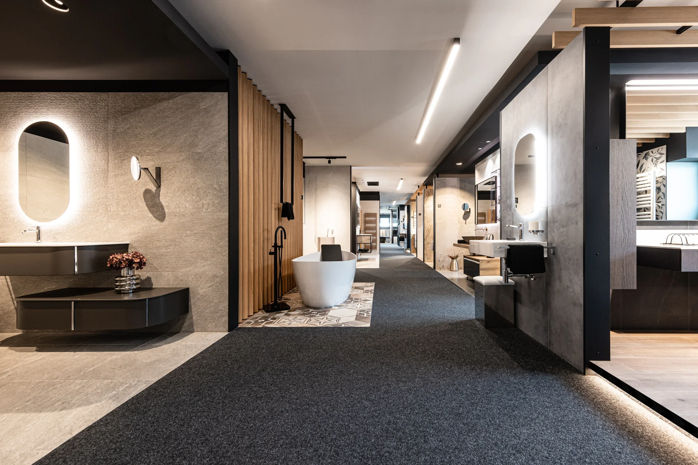 Ein modern eingerichteter Schauraum für Badezimmerdesign. Zu sehen sind verschiedene Bade- und Waschbereiche mit stilvollen Armaturen, Spiegeln und Holzverkleidungen. Ein freistehendes, weißes Badewannenmodell steht im Mittelpunkt, umgeben von elegant beleuchteten Waschplätzen. Der Raum wirkt geräumig und luxuriös.