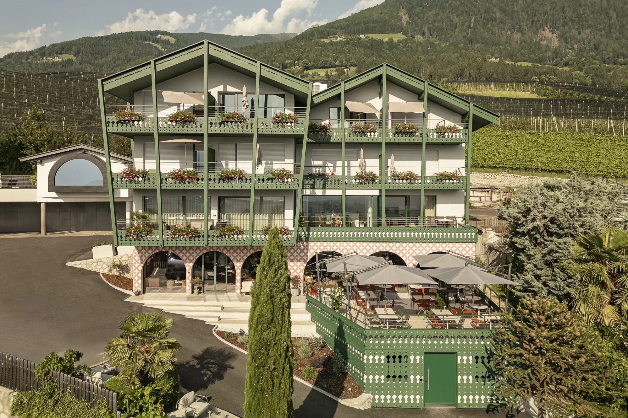 Ein dreistöckiges Hotel mit Balkonen, geschmückt mit Blumen, steht in einer grünen Berglandschaft. Eine Terrasse mit Tischen und Sonnenschirmen befindet sich im Vordergrund.