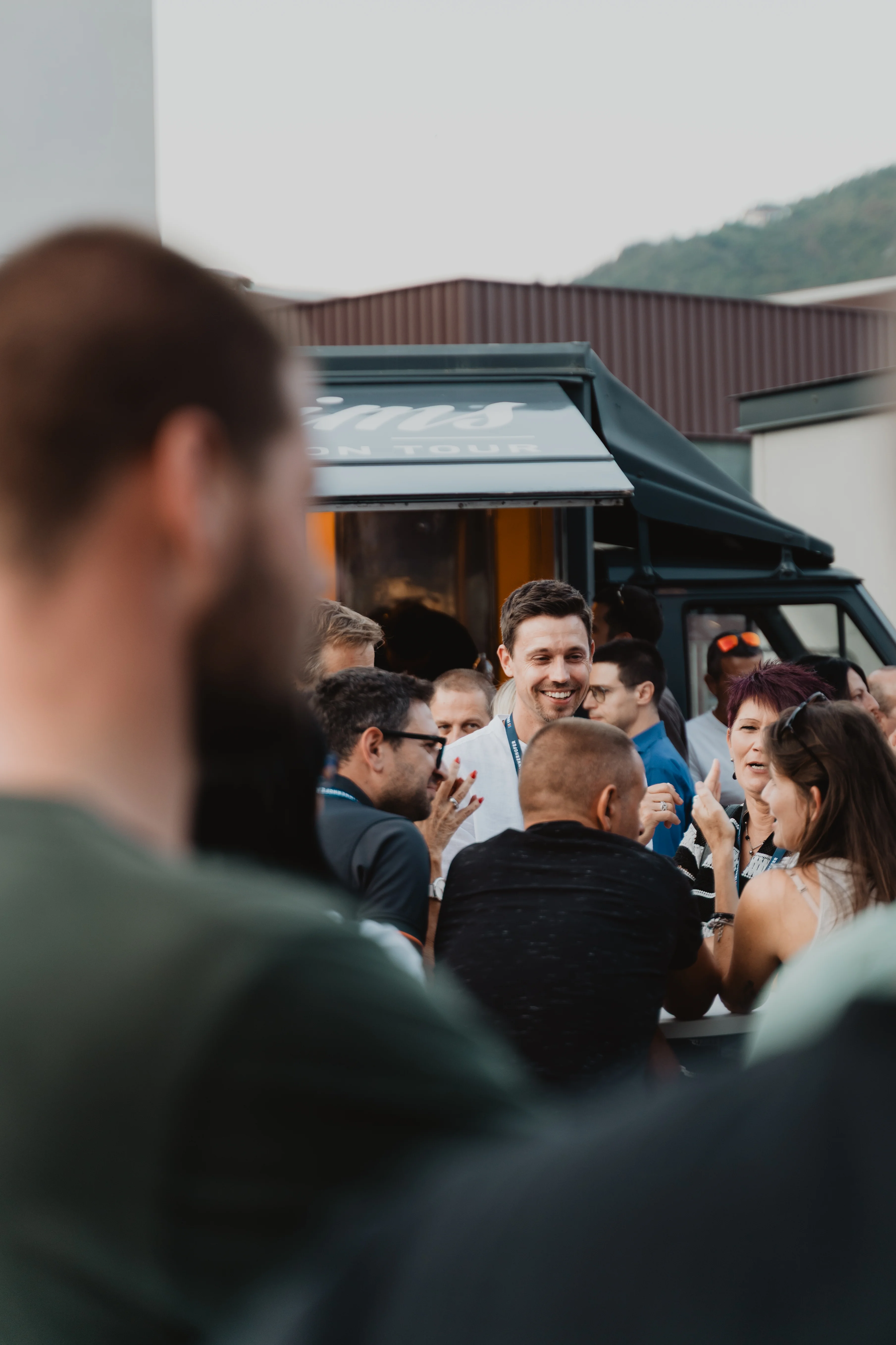 Eine Gruppe von Menschen unterhält sich und lacht vor einem schwarzen Foodtruck. Ein Mann im Mittelpunkt trägt ein weißes Hemd und lächelt. Im Hintergrund sind Gebäude und ein Hügel sichtbar.