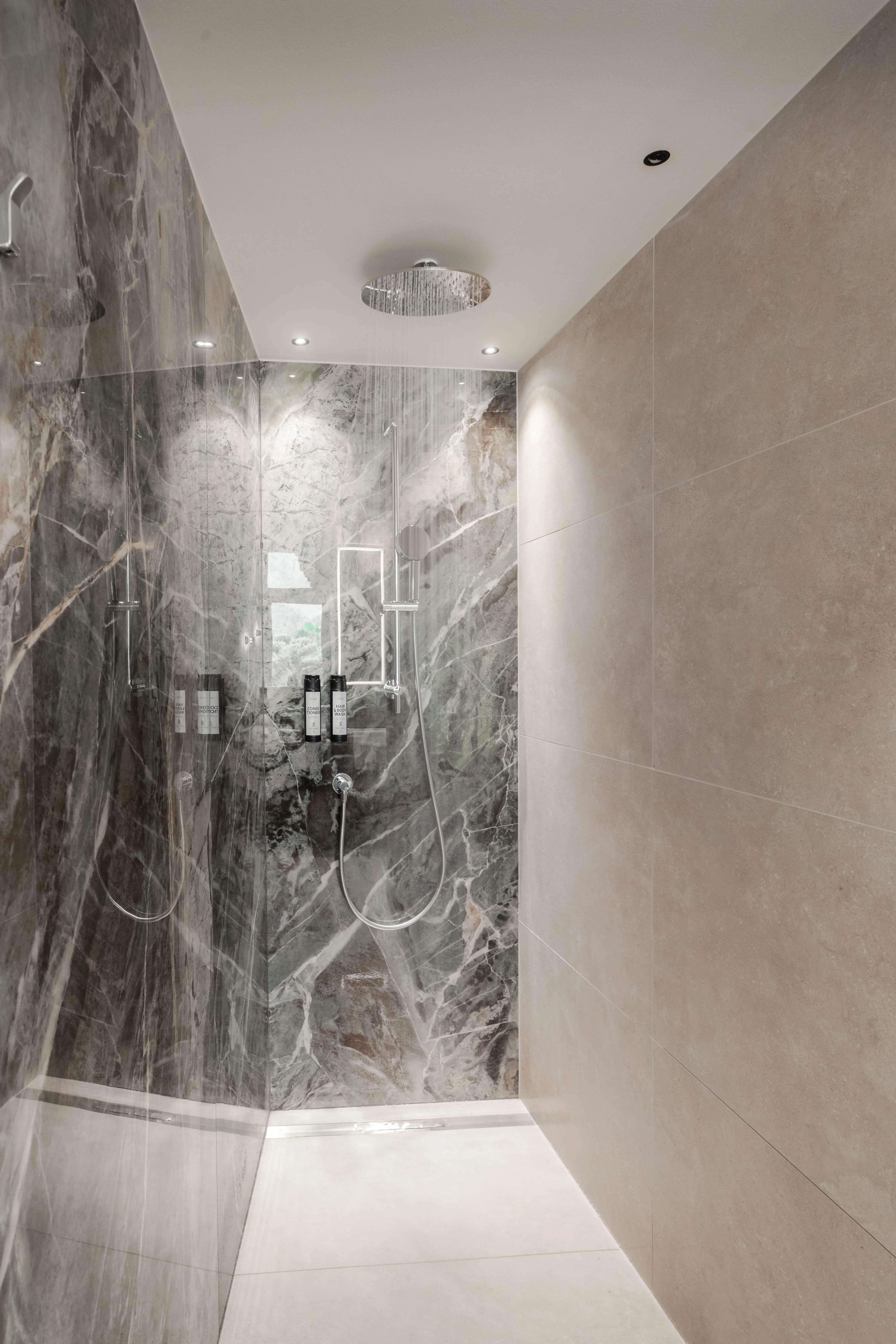 Luxuriöse, moderne Dusche mit großen grauen Marmorfliesen, Regenduschkopf an der Decke und eingebauter Duscharmatur. Die Beleuchtung ist dezent, aber effektiv, und es gibt eine harmonische Kombination aus Textur und Farbe.