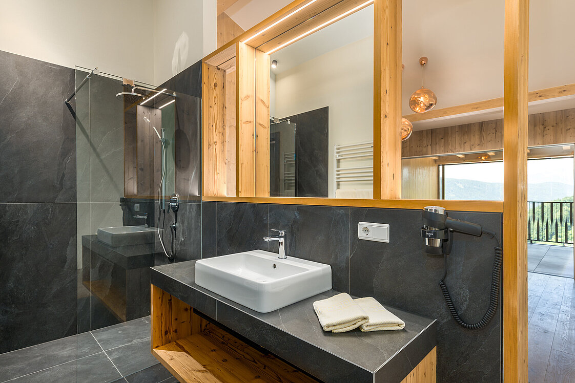 Bagno moderno con lavandino bianco su mensola in legno, specchio grande, doccia a vetro e pareti scure. Asciugacapelli montato a parete e due asciugamani piegati sul lavandino. Design elegante e minimalista.