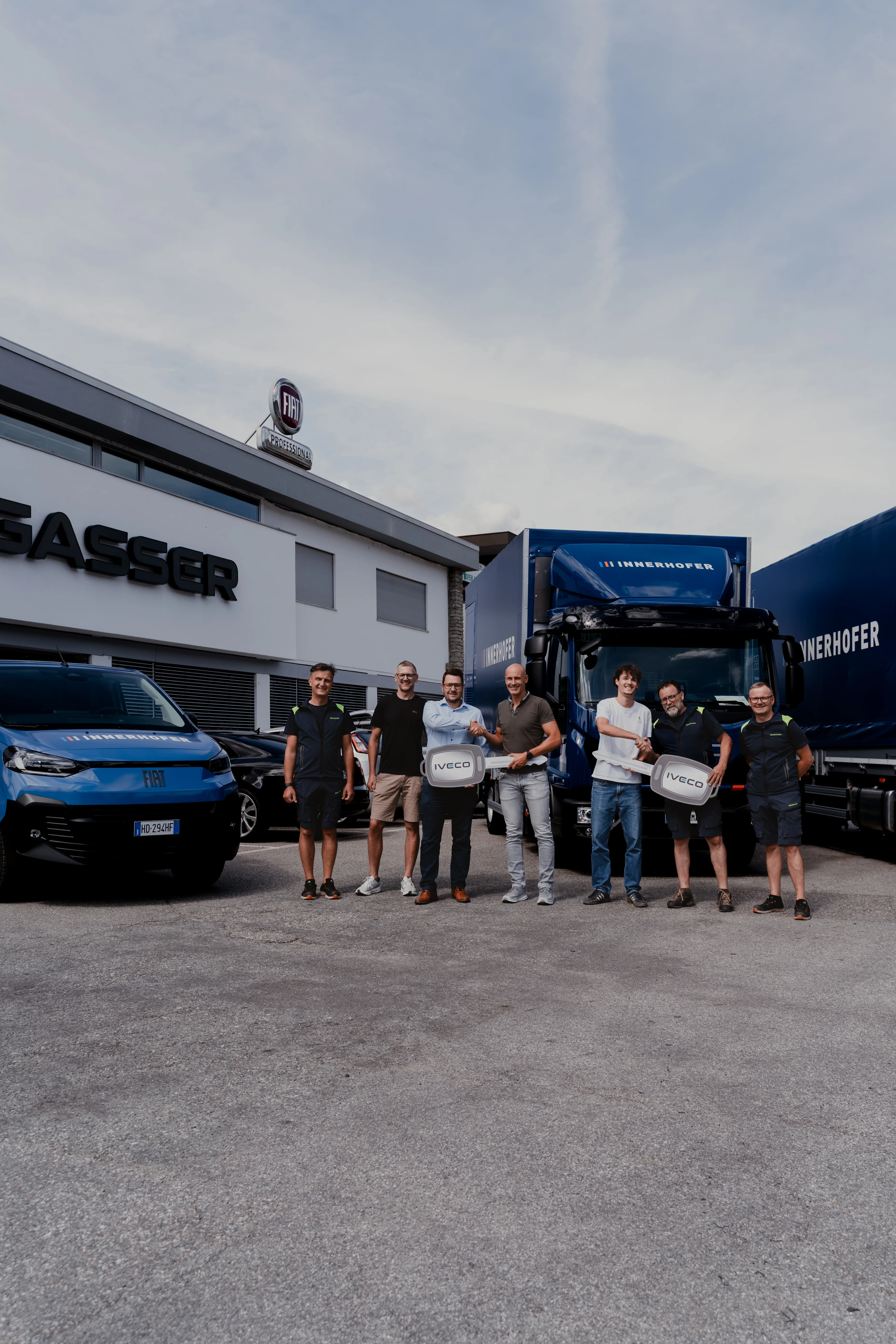 Eine Gruppe von sieben Personen steht vor zwei Lastwagen und einem Kleintransporter vor einem Gebäude mit der Aufschrift "R. LASSER". Sie halten große Fahrzeugschlüssel und lächeln in die Kamera. Der Himmel ist leicht bewölkt.