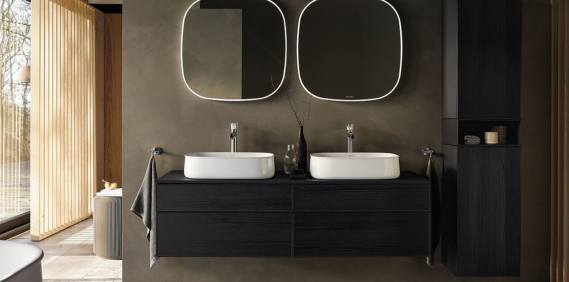 Modernes Badezimmer mit zwei Waschbecken, großen Spiegeln und Hängelampen. Akzente aus dunklem Holz und natürliches Licht durch ein großes Fenster auf der linken Seite.