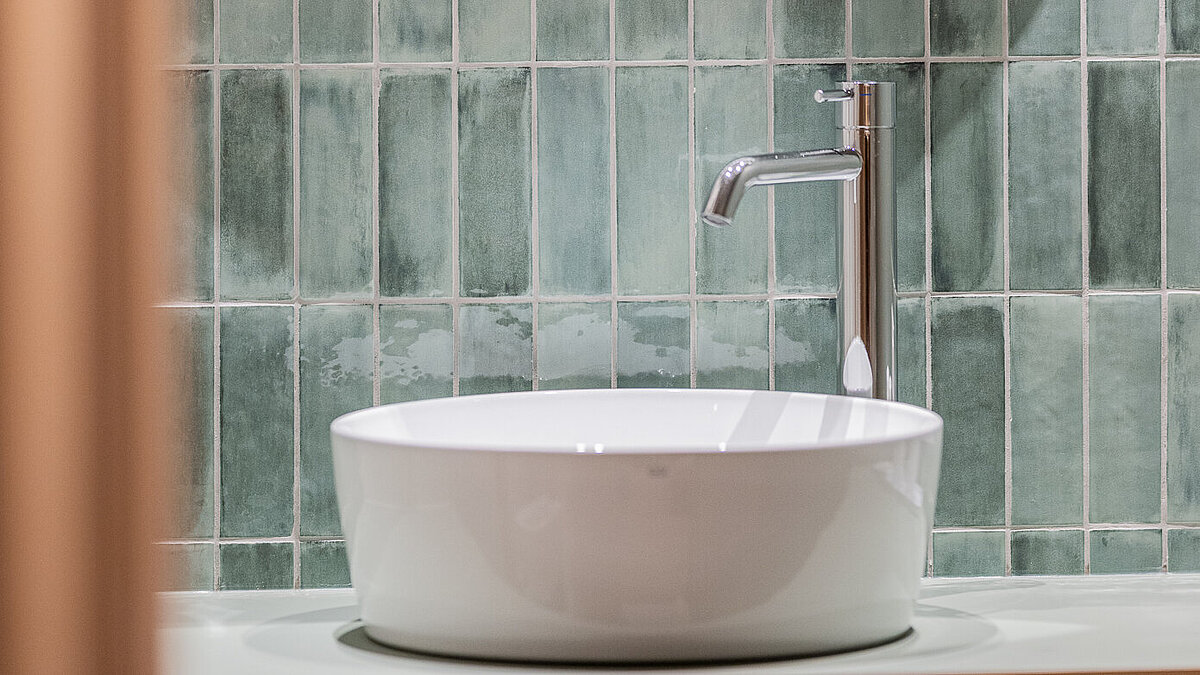 Ein rundes Waschbecken mit modernem Wasserhahn vor grünlichen Kacheln. Darüber hängt ein runder Spiegel. Das Design vermittelt Eleganz und Minimalismus.