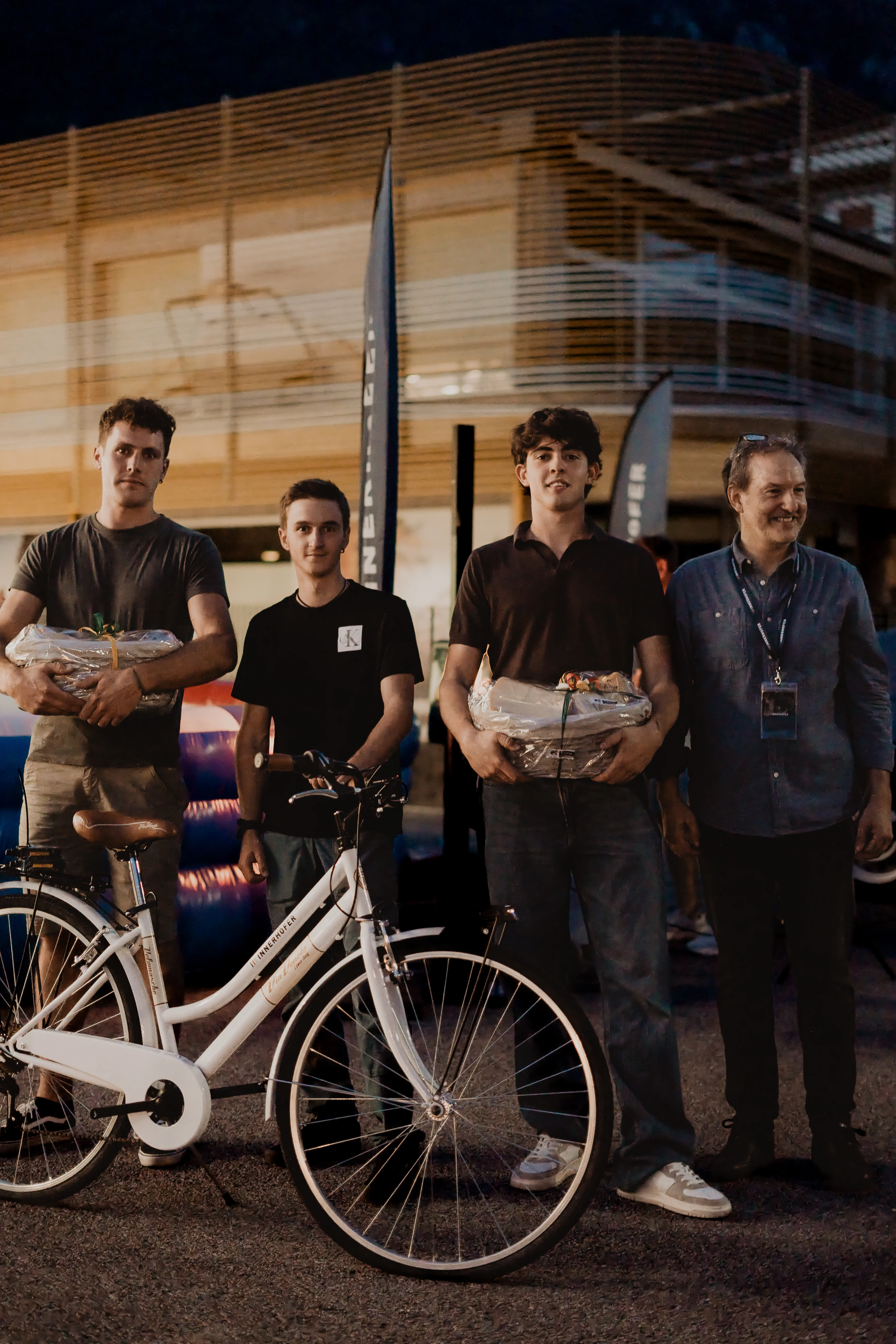 Vier Männer stehen nebeneinander vor einem modernen Gebäude. Drei von ihnen halten Geschenkkörbe in den Händen, während ein weißes Fahrrad vorne links im Bild steht. Sie tragen legere Kleidung und blicken leicht lächelnd in die Kamera.