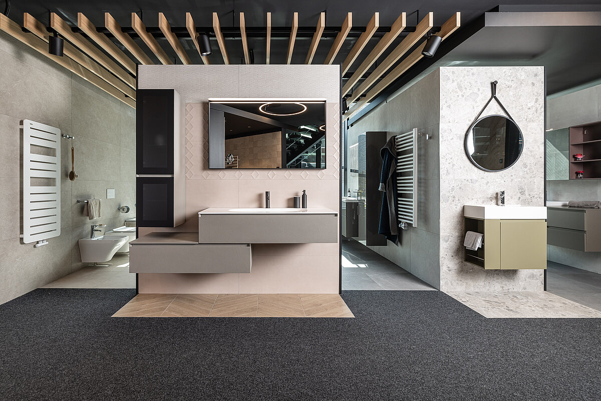 Modernes Badezimmerausstellungsraum mit Wandspiegeln, Waschbecken und einem mix aus offenen und geschlossenen Schränken. Die Raumdecke hat eine Holzbalkenoptik. Rechts befindet sich ein weiterer Waschbereich mit rundem Spiegel und kompakter Ablage.